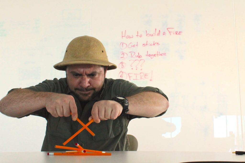 Man in a pith helmet trying to start a fire by rubbing two pencils together in front of a whiteboard.