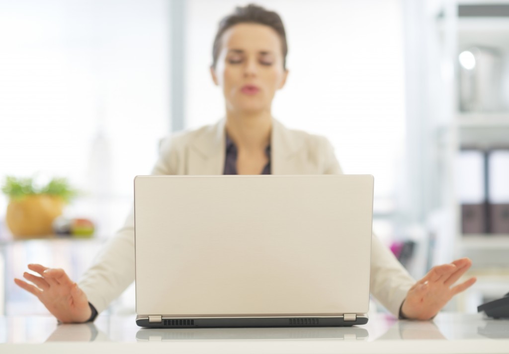 Photo of a person sitting behind a laptop with their hands to the side of the laptop and breathing out with their eyes closed.