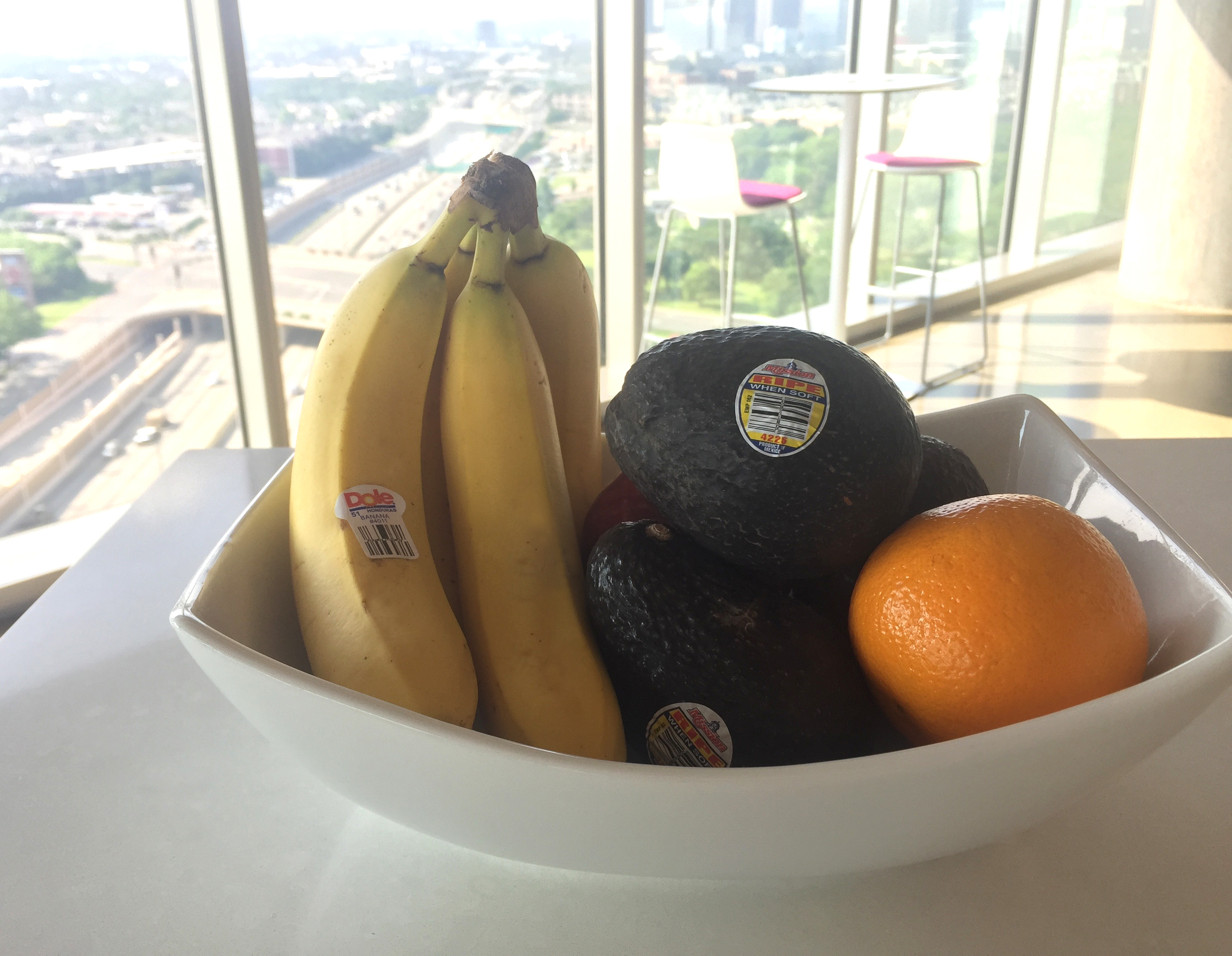Photo of a bowl of fruit containing bananas, avocados, and an orange with a highway scene in the background.