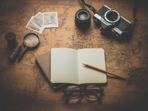 Photo of a magnifying glass, a journal, a pencil, a film camera, spectacles, and photos sitting on an old map.
