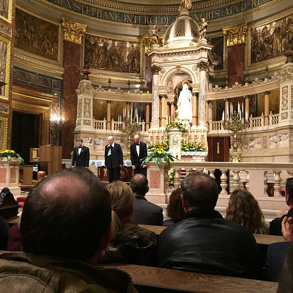 Men on stage in front of an audience for a concert in Budapest